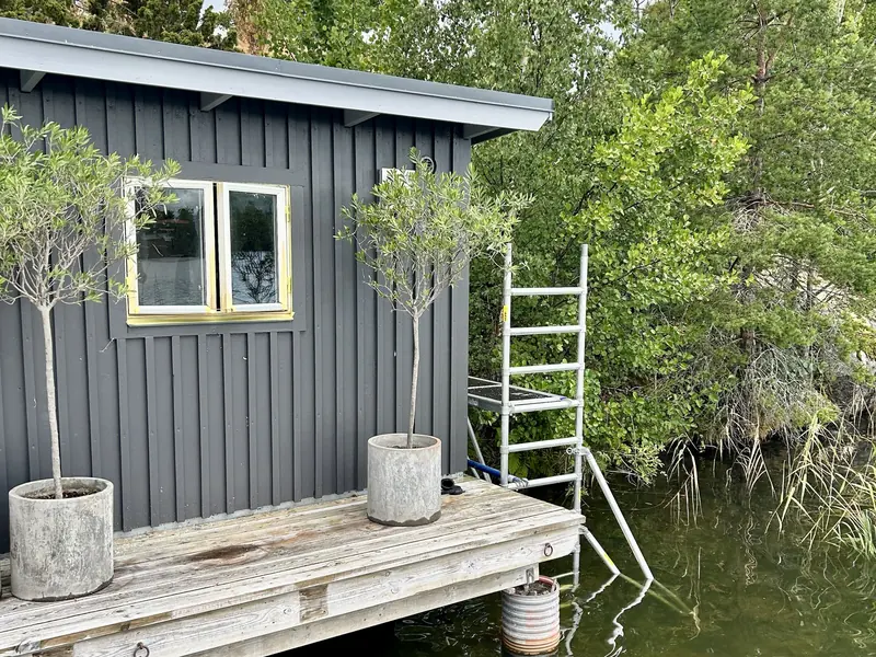 Sjöbodens fönster maskat inför målning, med fasaden färdigmålad och en byggställning placerad i vattnet vid gaveln